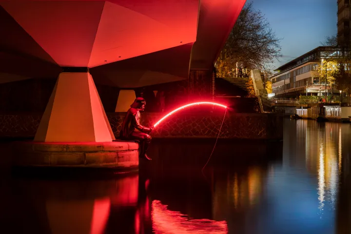 Person under bridge at night holding curved red light by water.