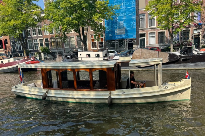 Small boat with flag on a canal, trees and buildings in background.