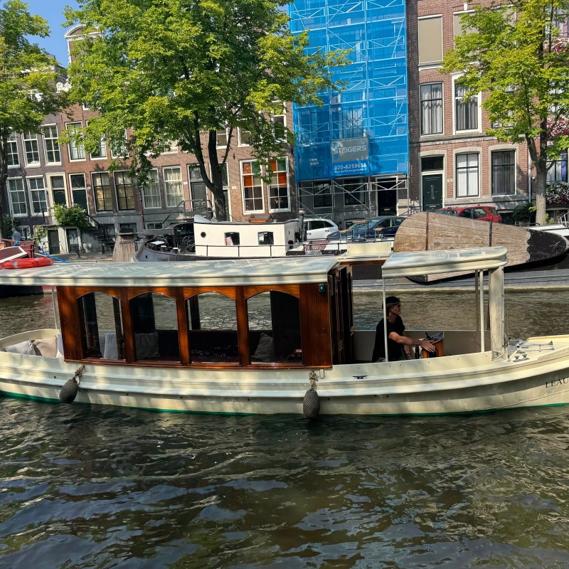 Small boat with flag on a canal, trees and buildings in background.