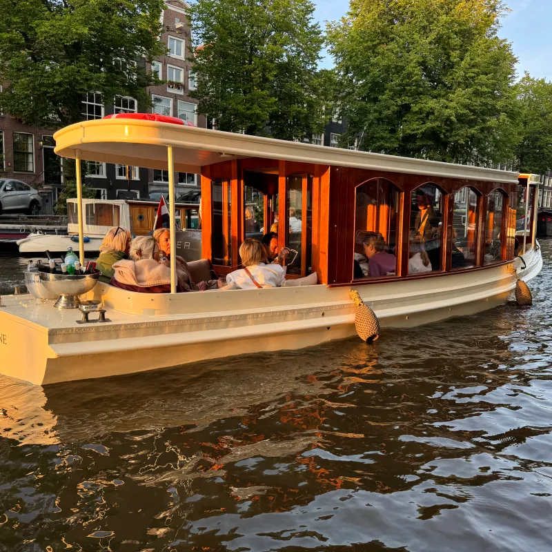 Calnal boat Leaudine for ride on a canal with Dutch flag, amidst trees and buildings in the background.